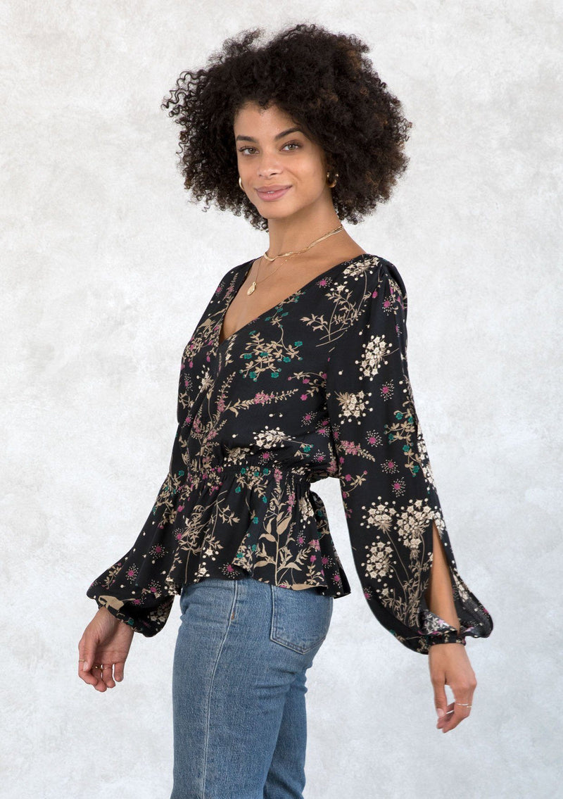 [Color: Black/Jade] A model wearing an ultra bohemian top in a black and green wildflower print. With voluminous long split sleeves, a peplum waist, and an open back with tassel tie closure.