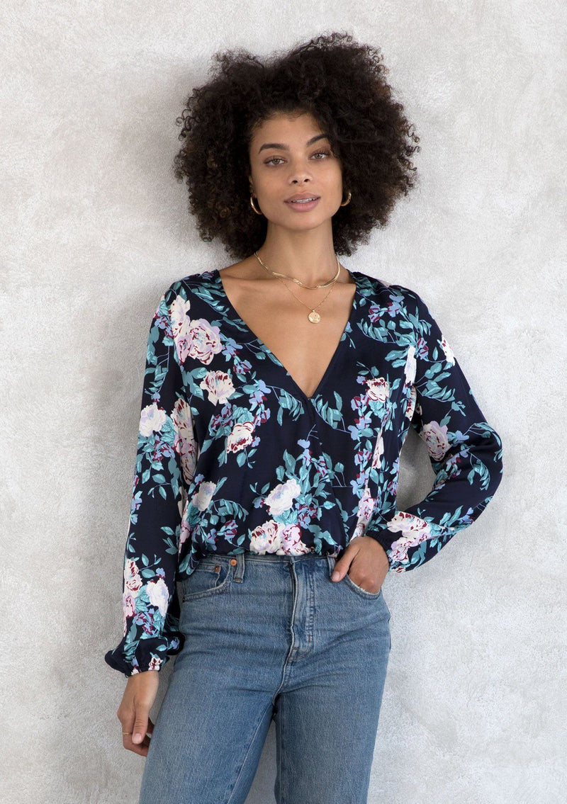 [Color: Navy/Lilac] A model wearing a navy and lilac floral print silky blouse. With a surplice neckline in the front and back, a smocked elastic hemline, and long voluminous sleeves. 