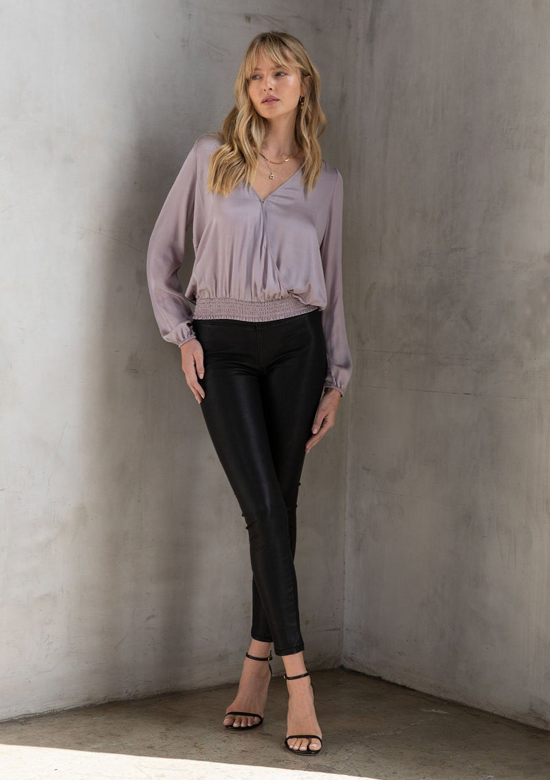 [Color: Lilac Mist] A model wearing a silky bohemian top, in lilac purple. With a surplice v neckline in front and back and a smocked elastic hemline. An effortless bohemian holiday blouse.
