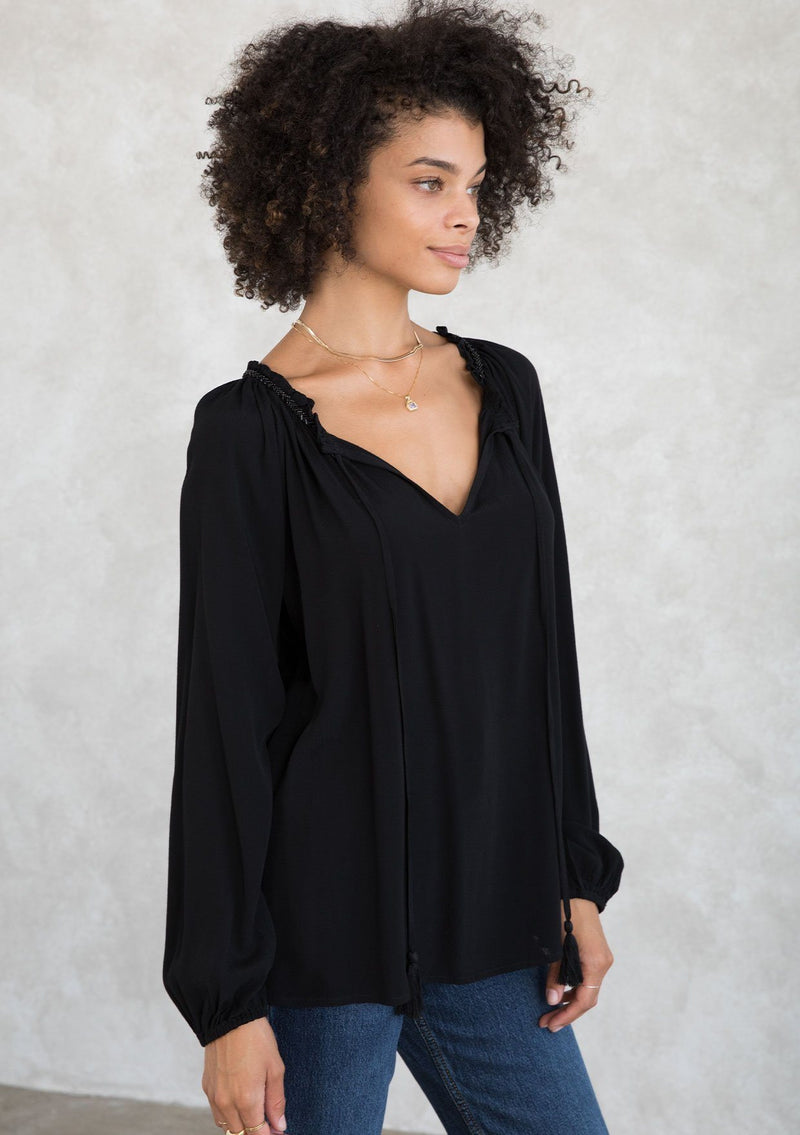 [Color: Black] A model wearing a Holiday ready, classic black peasant top in textured crepe, with beaded trim along the neckline. With a split neckline and tassel ties. A timeless blouse for your next special occasion. 