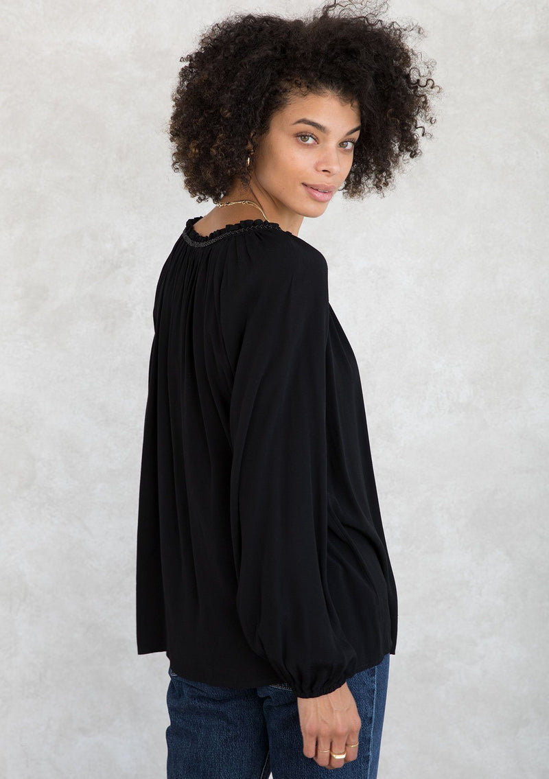 [Color: Black] A model wearing a Holiday ready, classic black peasant top in textured crepe, with beaded trim along the neckline. With a split neckline and tassel ties. A timeless blouse for your next special occasion. 
