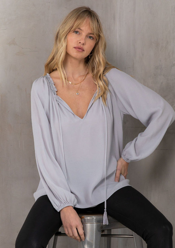 [Color: Platinum] A model wearing a Holiday ready, classic grey peasant top in textured crepe, with beaded trim along the neckline. With a split neckline and tassel ties. A timeless blouse for your next special occasion. 