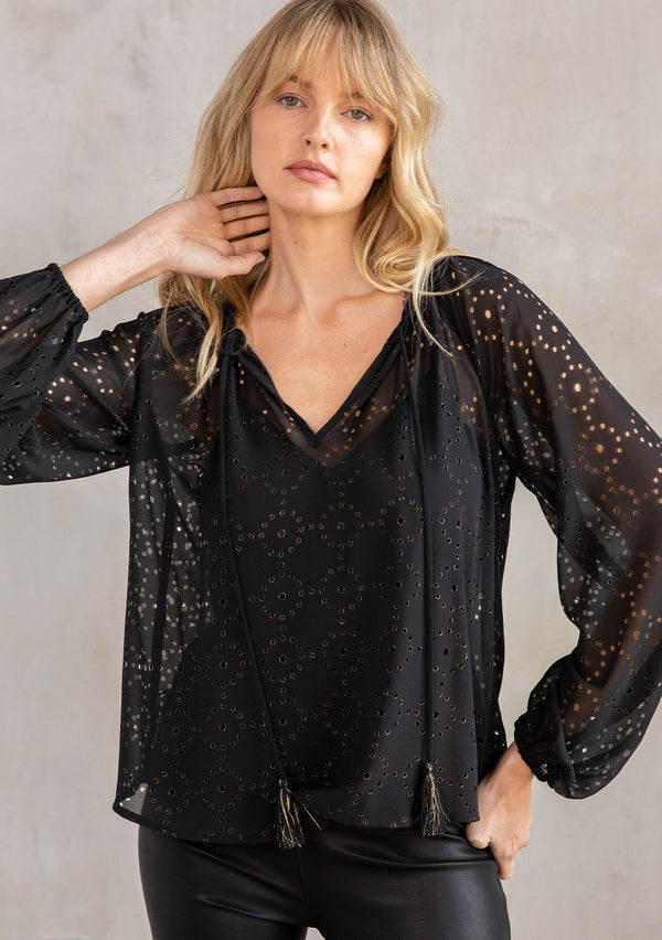[Color: Black] A model wearing a dreamy sheer chiffon black peasant top with gold laser cut out details. With a lace trimmed neckline, long sleeves, and tassel ties. 