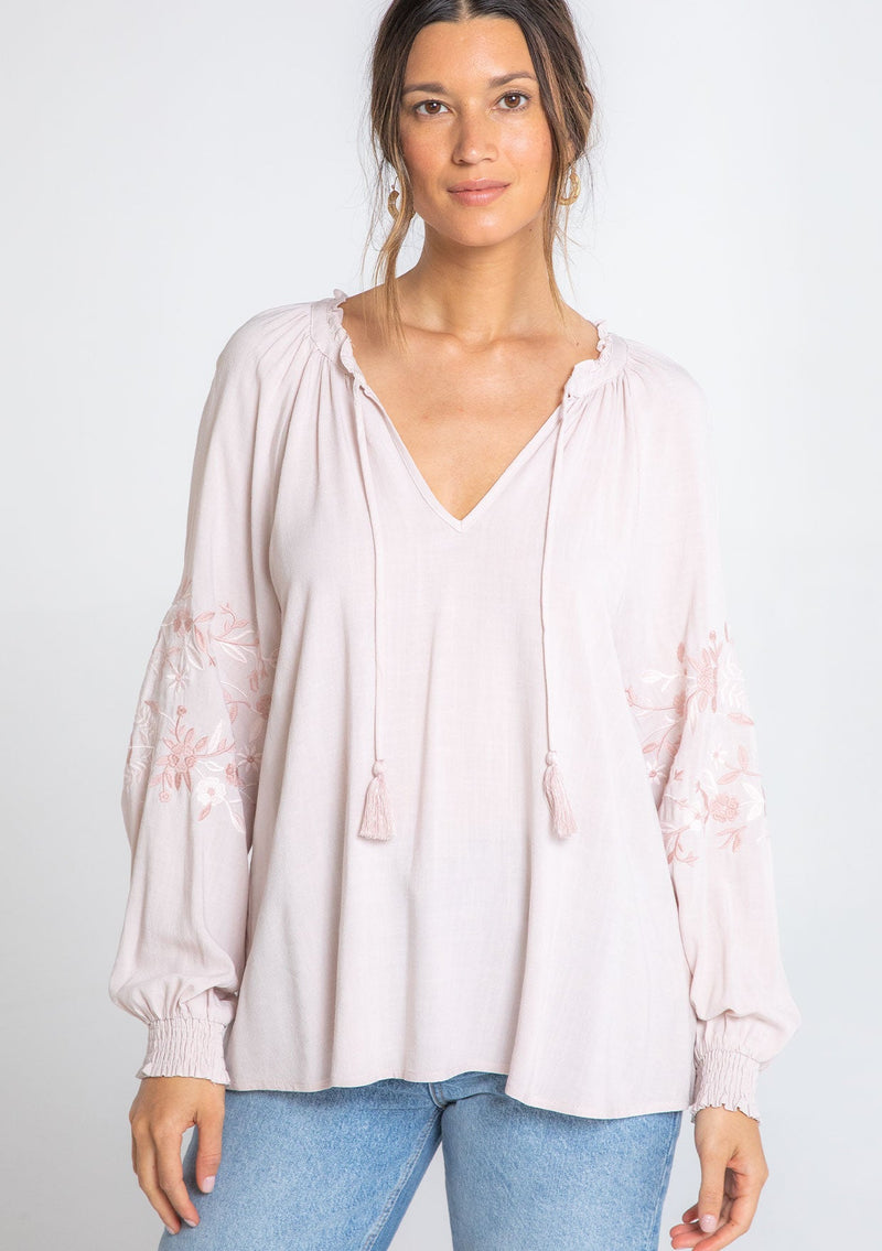 [Color: Blush/Rose] A model wearing a flowy bohemian pink peasant top with voluminous long sleeves, embroidered sleeve details, and a tassel tie v neckline. 