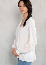 [Color: Vanilla/Sand] A model wearing a flowy bohemian off white peasant top with voluminous long sleeves, embroidered sleeve details, and a tassel tie v neckline. 