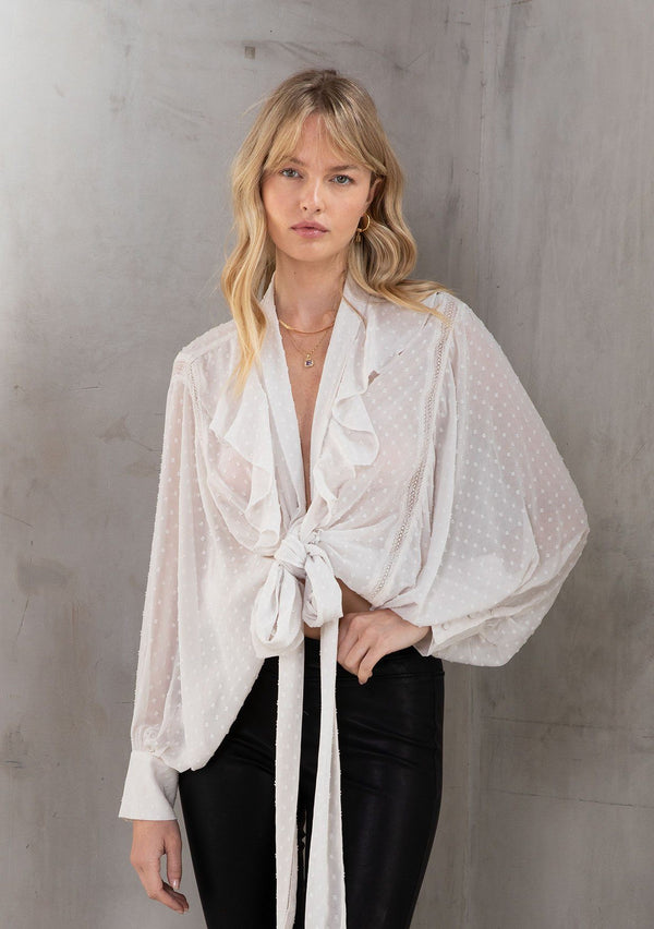 [Color: Gardenia] A model wearing a sheer swiss dot ivory bohemian top. With a ruffled lapel, long batwing sleeves, a self covered button cuff wrist, and a tie front detail that can be tied in multiple ways.