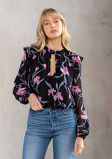 [Color: Black/Lilac] A model wearing an ultra pretty sheer floral blouse in black and lilac purple clip dot chiffon. With a front keyhole, long sheer sleeves, and a smocked high neckline. 