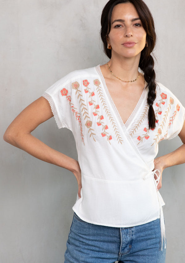 [Color: Ivory/Coral] A model wearing a white bohemian wrap top with floral embroidery detail, lace trim, and short sleeves. 