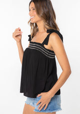 [Color: Black] A woman wearing a black crinkle gauze flowy bohemian tie shoulder tank top with smocked neckline. 