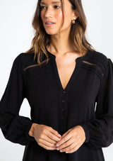 [Color: Black] A model wearing a timeless long sleeve black linen blend blouse. With a decorative button front, delicate lattice trim, and pleated details.