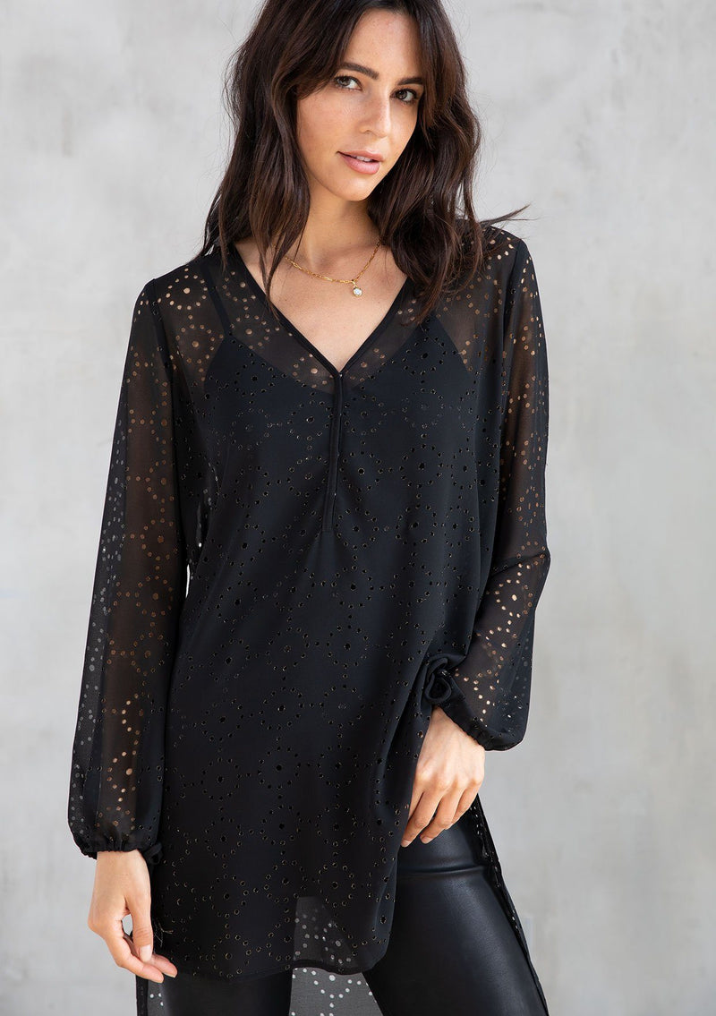 [Color: Black] A model wearing a black chiffon tunic top with gold metallic laser cut details throughout. With tassel tie wrist cuffs, a high low hemline, and a snap button front. 
