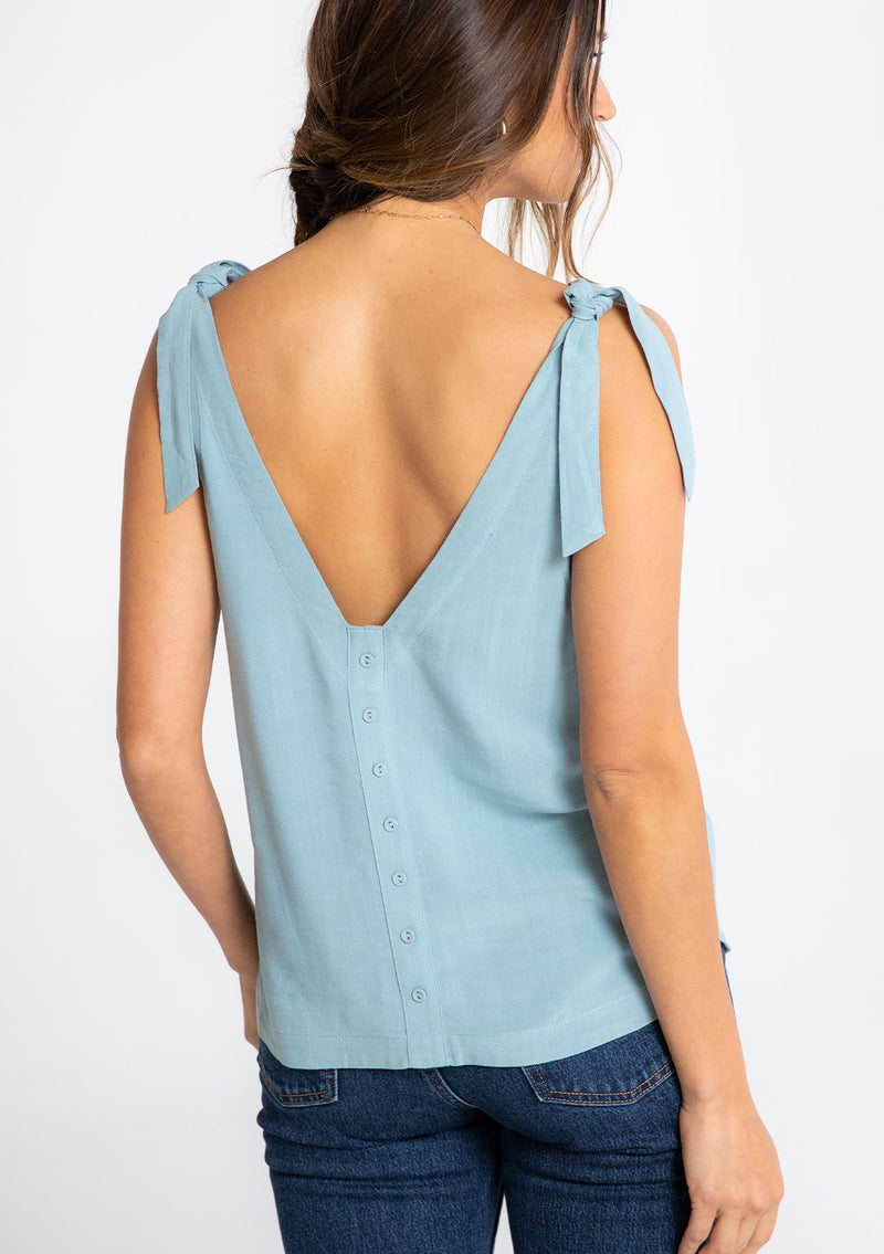 [Color: Dusty Teal] A model wearing a teal linen blend tank top with a tie shoulder and a button up back detail. 