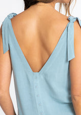 [Color: Dusty Teal] A model wearing a teal linen blend tank top with a tie shoulder and a button up back detail. 