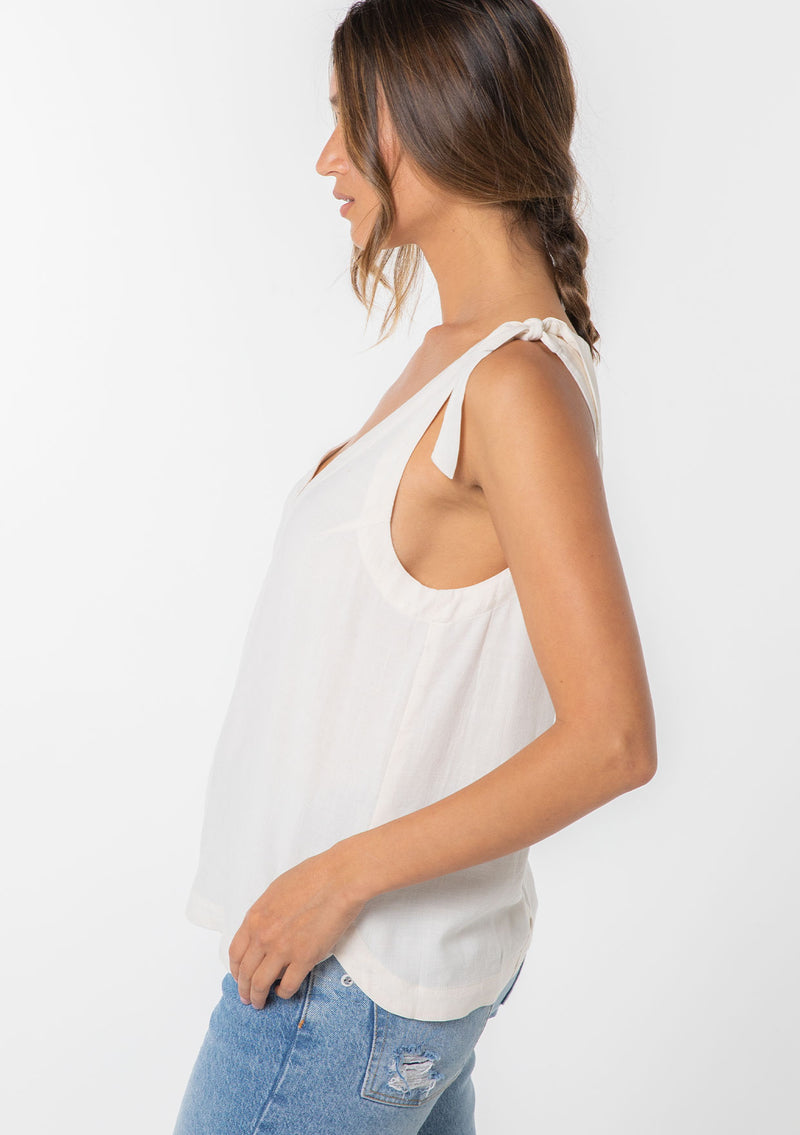[Color: Vanilla] A model wearing an off white linen blend tank top with a tie shoulder and a button up back detail. 