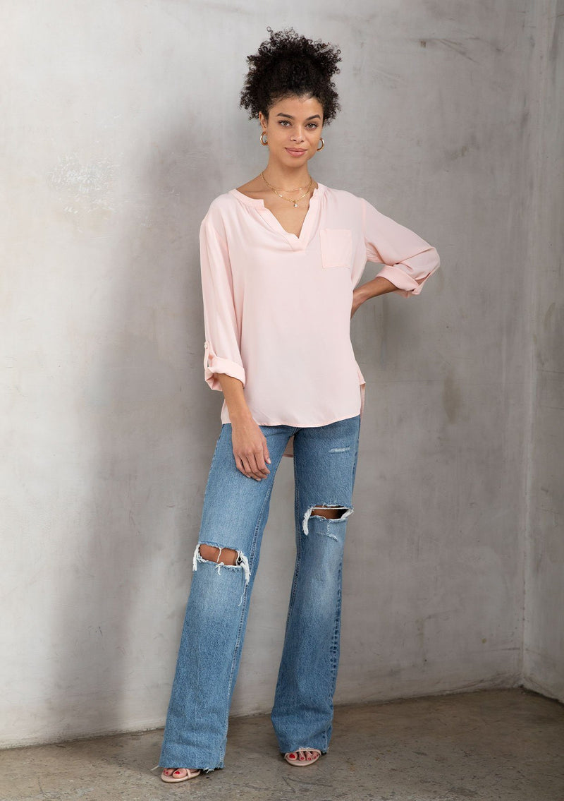 [Color: Bellini] A model wearing a silky light pink blouse with rolled tab sleeves, a front patch pocket, and a split v neckline. Classic top for the office.