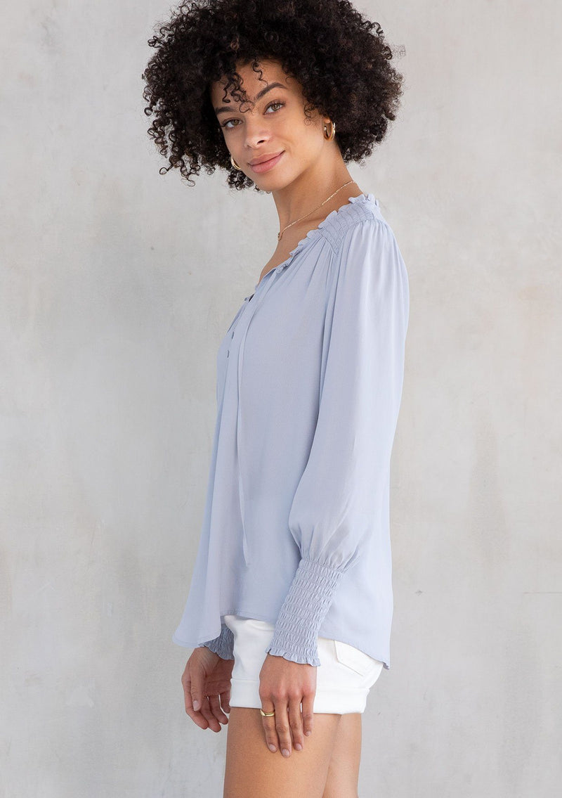 [Color: Powder Blue] A model wearing a classic light blue peasant top, designed in silky crepe. Featuring a split v neckline with ties and long voluminous sleeves.