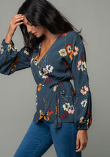 [Color: Grey/Wine] A side facing image of a brunette model wearing a slate blue grey long sleeve wrap top with a wine red floral print. 