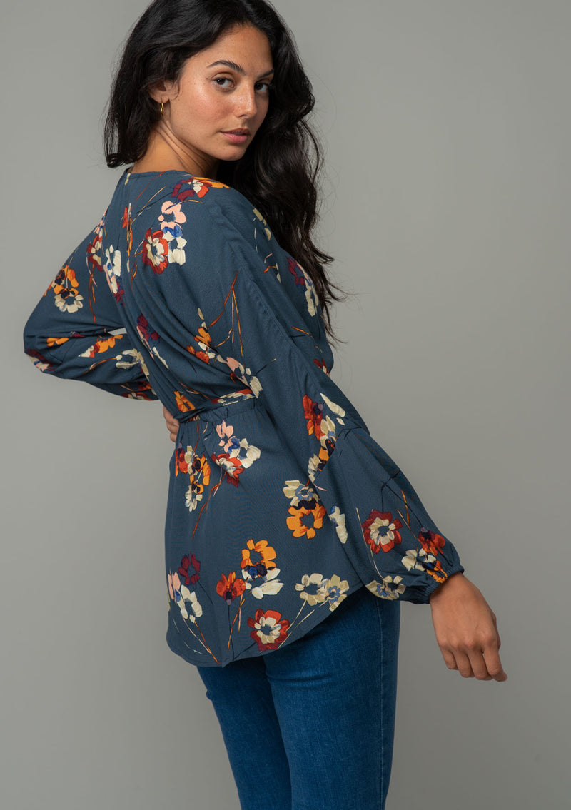 [Color: Grey/Wine] A back facing image of a brunette model wearing a slate blue grey long sleeve wrap top with a wine red floral print. The model is looking over her shoulder. 