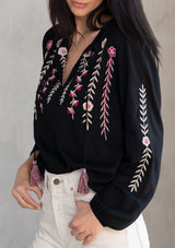[Color: Black/Mulberry] A model wearing a black flowy bohemian peasant top with floral embroidery, tassel neck ties, and voluminous long sleeves. 