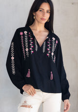 [Color: Black/Mulberry] A model wearing a black flowy bohemian peasant top with floral embroidery, tassel neck ties, and voluminous long sleeves. 
