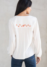 [Color: Ivory/Coral] A model wearing a white flowy bohemian peasant top with floral embroidery, tassel neck ties, and voluminous long sleeves. 