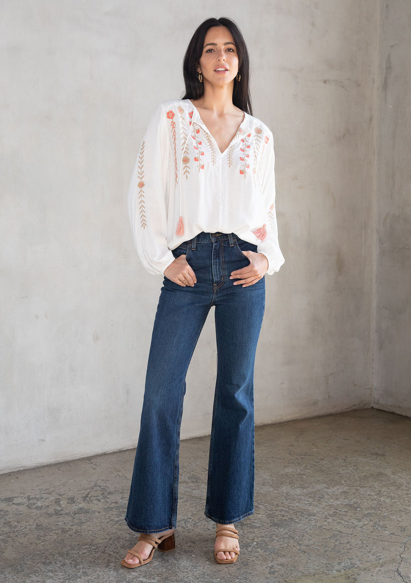 [Color: Ivory/Coral] A model wearing a white flowy bohemian peasant top with floral embroidery, tassel neck ties, and voluminous long sleeves. 