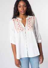 [Color: Ivory/Coral] A model wearing a soft white bohemian button front shirt with long rolled sleeves and floral embroidered details.