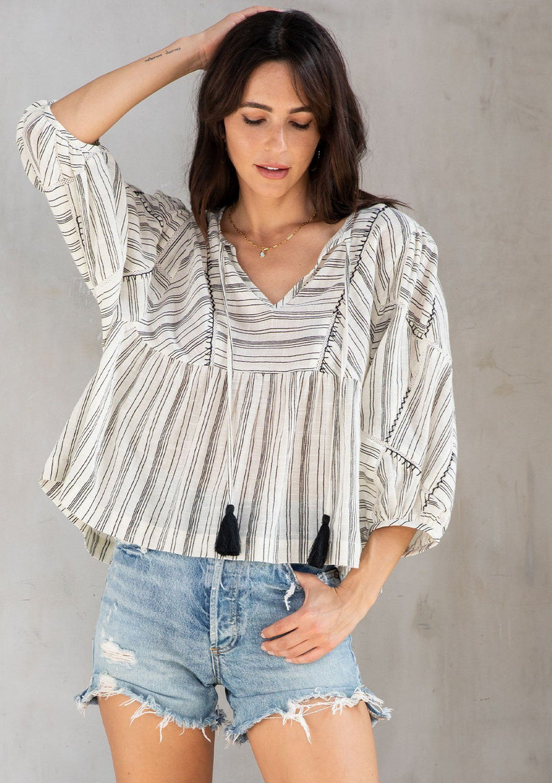 [Color: Natural/Black] A model wearing an off white cotton bohemian blouse in a black stripe with hand stitched embroidered details. With patchwork puff sleeves and a split neckline with tassel ties. 