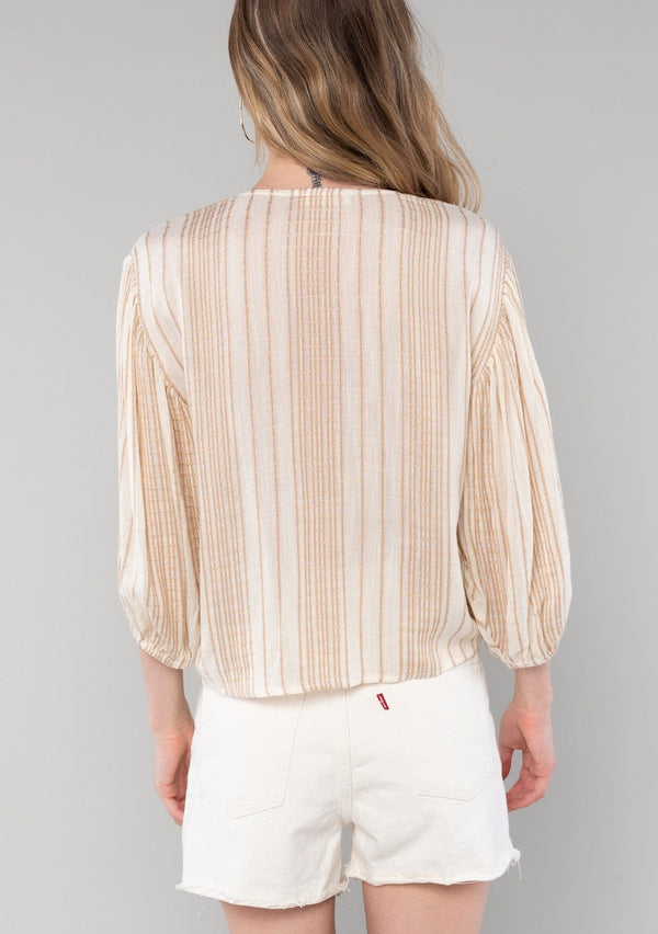 [Color: Natural/Tan] A back facing image of a blonde model wearing a resort ready bohemian top in a natural and tan stripe. With long sleeves, a v neckline, and a tie front waist. 