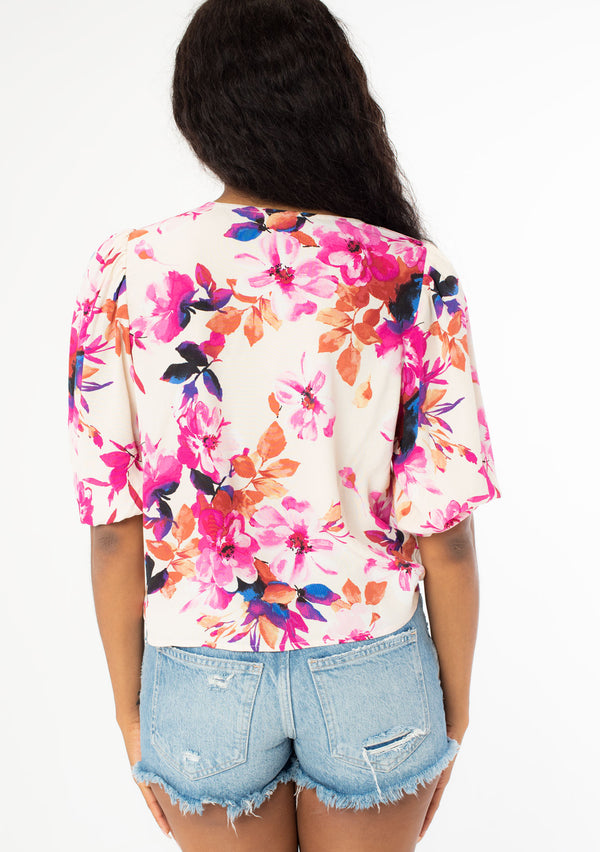 [Color: Ivory/Fuchsia] A back facing image of a black model wearing a pink and white watercolor floral print top with a tie front waist detail, half length sleeves, and a v neckline. 