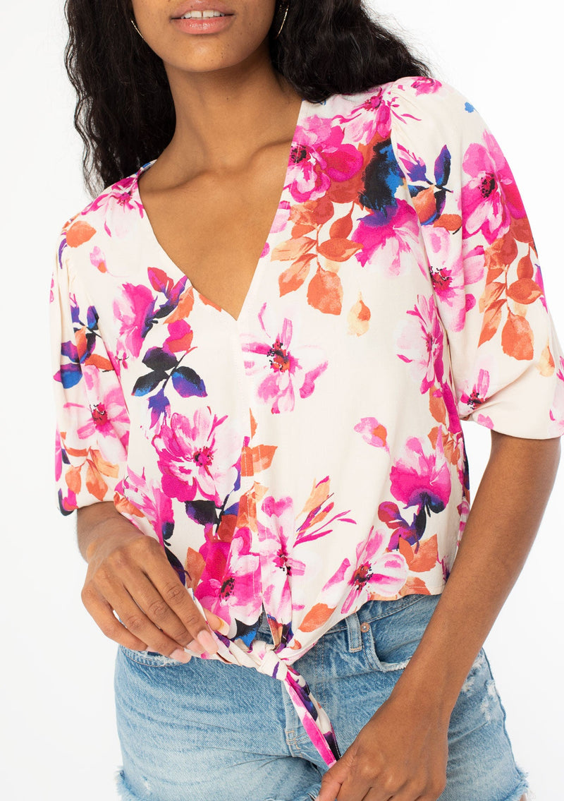 [Color: Ivory/Fuchsia] A close up front facing image of a black model wearing a pink and white watercolor floral print top with a tie front waist detail, half length sleeves, and a v neckline. 