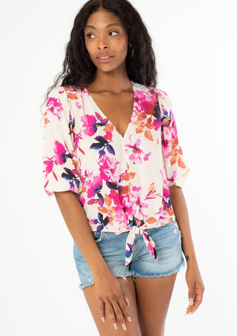 [Color: Ivory/Fuchsia] A front facing image of a black model wearing a pink and white watercolor floral print top with a tie front waist detail, half length sleeves, and a v neckline. 