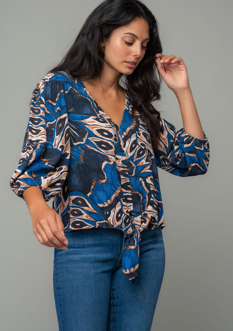 [Color: Cobalt/Tan] A half body front facing image of a brunette model wearing a navy blue and tan butterfly wing print top with three quarter length voluminous sleeves and a tie front detail. 