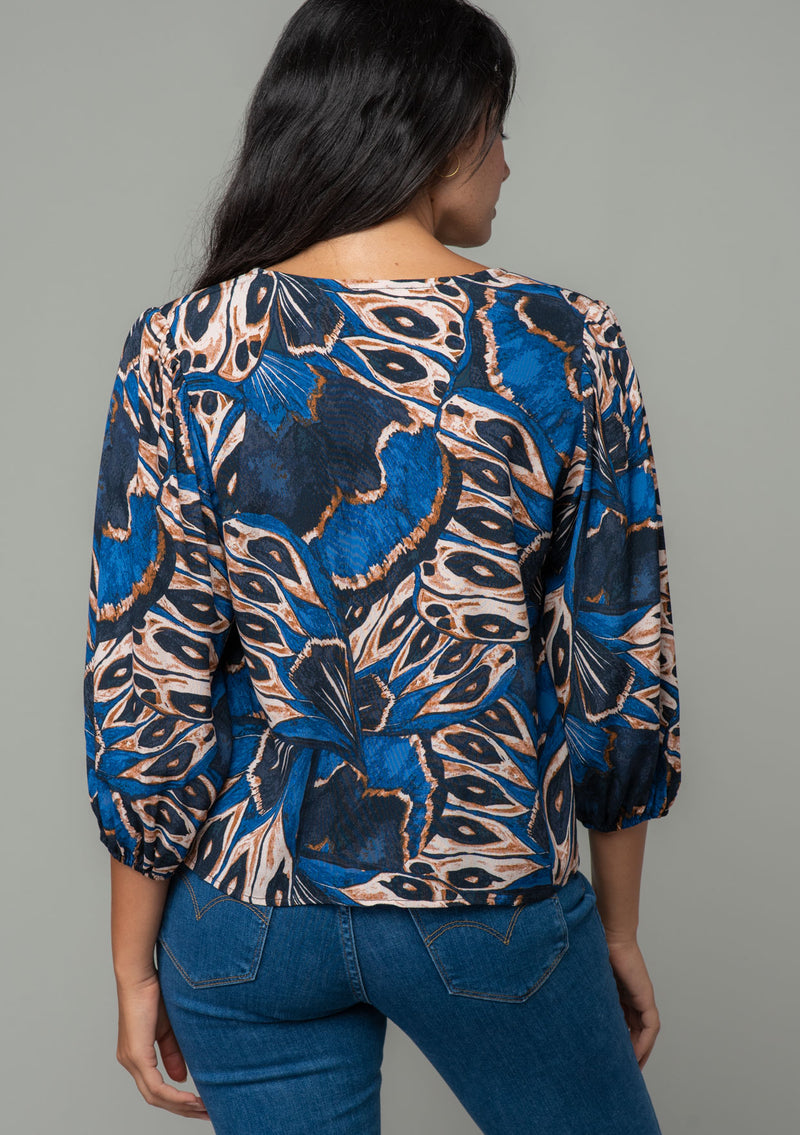 [Color: Cobalt/Tan] A back facing image of a brunette model wearing a navy blue and tan butterfly wing print top with three quarter length voluminous sleeves and a tie front detail. 