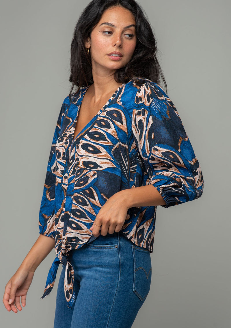 [Color: Cobalt/Tan] A side facing image of a brunette model wearing a navy blue and tan butterfly wing print top with three quarter length voluminous sleeves and a tie front detail. 
