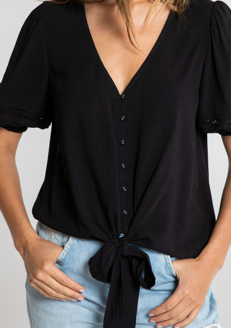 [Color: Black] A model wearing a timeless black linen blend bohemian top. With short puff sleeves, an adjustable tie front waist detail, and a button front.