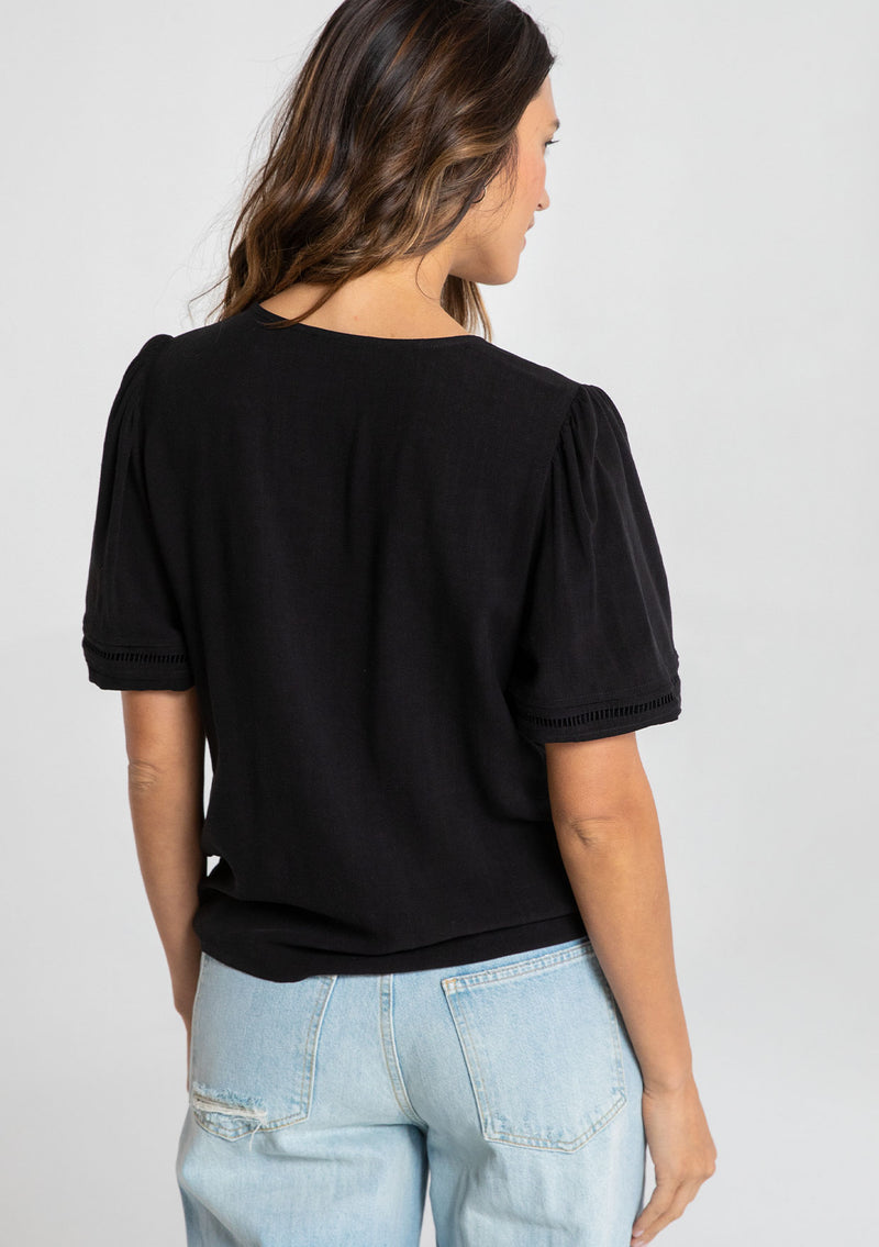 [Color: Black] A model wearing a timeless black linen blend bohemian top. With short puff sleeves, an adjustable tie front waist detail, and a button front.