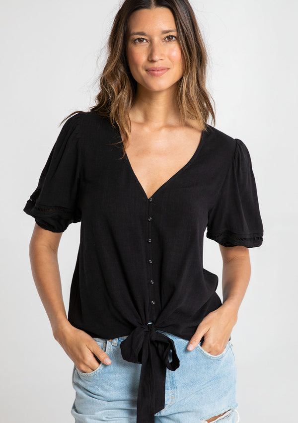 [Color: Black] A model wearing a timeless black linen blend bohemian top. With short puff sleeves, an adjustable tie front waist detail, and a button front.