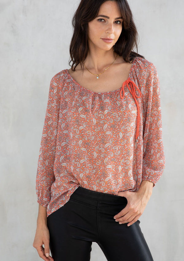 [Color: Coral/Natural] A model wearing a delicate sheer chiffon blouse in a coral paisley print with gold lurex detail. Featuring a tassel tie drawstring wide neckline. 