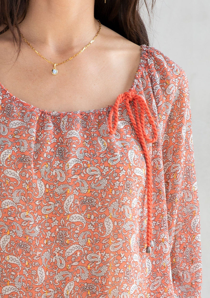 [Color: Coral/Natural] A model wearing a delicate sheer chiffon blouse in a coral paisley print with gold lurex detail. Featuring a tassel tie drawstring wide neckline. 