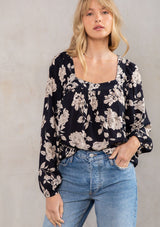 [Color: Black/Natural] A model wearing a flowy bohemian blouse in a black and natural floral print. With long voluminous sleeves, a trendy square neckline, and adjustable tie back detail.