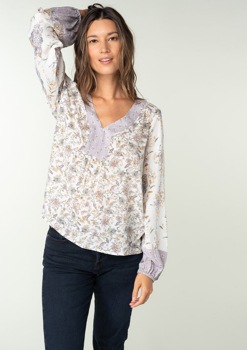 [Color: Natural/Dusty Lilac] A half body front facing image of a brunette model wearing a bohemian blouse in a lilac purple and natural off white mixed floral print. With voluminous long sleeves and a v neckline.