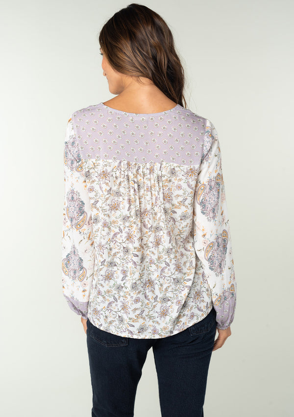 [Color: Natural/Dusty Lilac] A back facing image of a brunette model wearing a bohemian blouse in a lilac purple and natural off white mixed floral print. With voluminous long sleeves and a v neckline. 