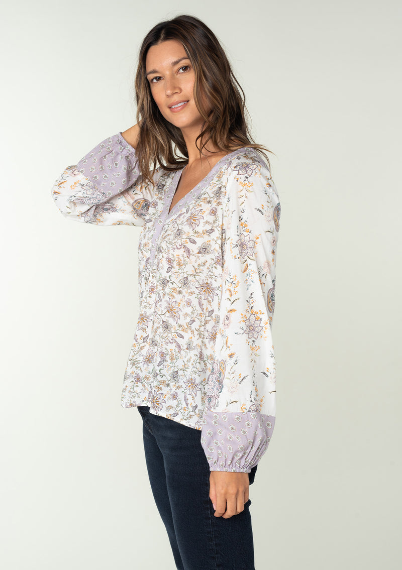 [Color: Natural/Dusty Lilac] A side facing image of a brunette model wearing a bohemian blouse in a lilac purple and natural off white mixed floral print. With voluminous long sleeves and a v neckline.