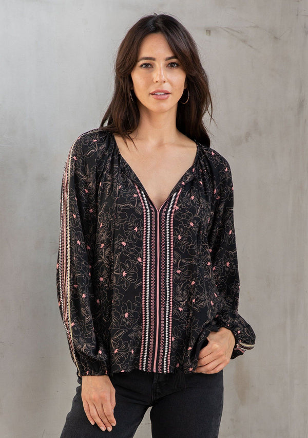 [Color: Black/Rose] A model wearing a classic black bohemian peasant top with romantic pink floral print. With long voluminous sleeves and a split v neckline with tassel ties. 