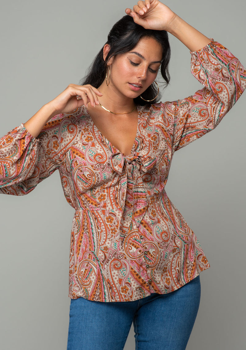 [Color: Natural/Rust] A front facing image of a brunette model wearing a bohemian blouse in a natural and rust red paisley print. With long sleeves, a peplum waist, and a v neckline with tie front detail. 