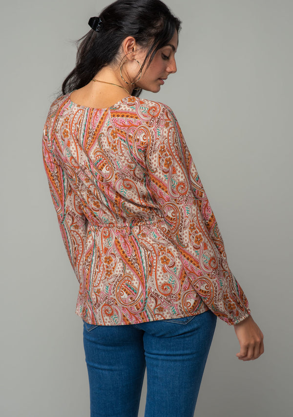 [Color: Natural/Rust] A back facing image of a brunette model wearing a bohemian blouse in a natural and rust red paisley print. With long sleeves, a peplum waist, and a v neckline with tie front detail. 