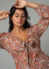 [Color: Natural/Rust] A close up front facing image of a brunette model wearing a bohemian blouse in a natural and rust red paisley print. With long sleeves, a peplum waist, and a v neckline with tie front detail. 