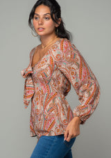 [Color: Natural/Rust] A side facing image of a brunette model wearing a bohemian blouse in a natural and rust red paisley print. With long sleeves, a peplum waist, and a v neckline with tie front detail. 