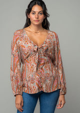 [Color: Natural/Rust] A half body front facing image of a brunette model wearing a bohemian blouse in a natural and rust red paisley print. With long sleeves, a peplum waist, and a v neckline with tie front detail. 